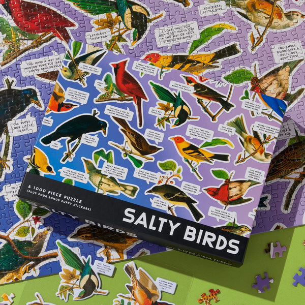 A Brass Monkey Salty Birds 1000 Piece Puzzle box sits atop a half-finished jigsaw, both featuring colorful illustrated birds and witty, salty captions.