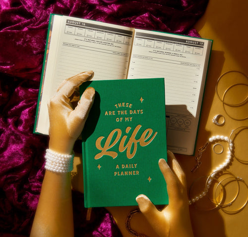 A person with pearl bracelets and gold gloves holds the Brass Monkey "These are the Days of My Life Undated Planner," while an open planner and gold jewelry sit on a golden surface with purple fabric in the background.