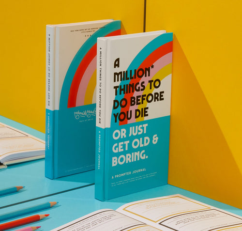 The Brass Monkey A Million Things to Do Before You Die Prompted Journal stands upright on a yellow surface, surrounded by open journals and colored pencils.