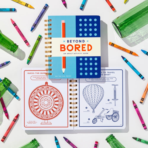 The spiral-bound “Beyond Bored” activity book by Brass Monkey is open to adult puzzles, including a guess-the-number game and hot air balloon drawing, surrounded by colorful crayons and green glass bottles.