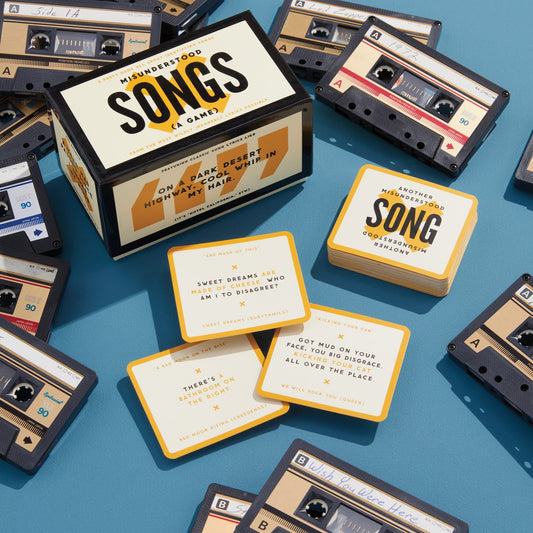 The Brass Monkey Misunderstood Songs Game is shown, featuring cassette tape-shaped cards and yellow question cards with misheard lyrics from hit songs, all displayed on a blue surface with the game box also visible.
