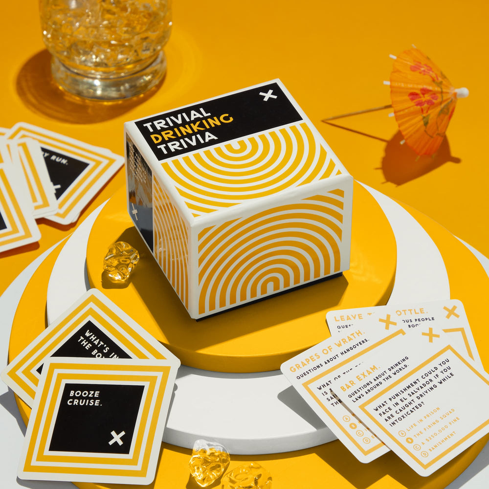 A box of Brass Monkey's Trivial Drinking Trivia sits on stacked white and yellow plates, surrounded by party cards, ice cubes, a drink in a glass, and a yellow cocktail umbrella—all set against a bright yellow background.